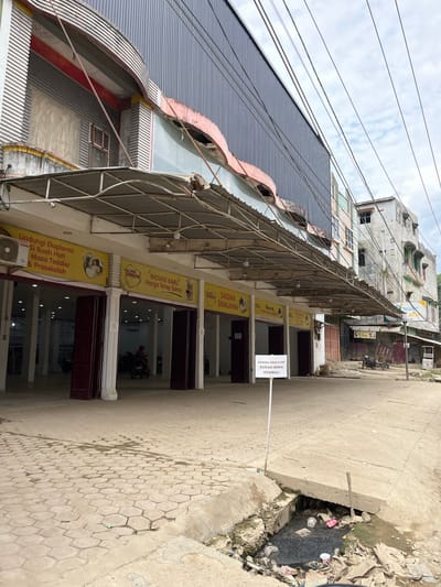 The image captures a street scene in Kota Kuala Simpang, Indonesia, focusing on the exterior of a commercial building with several storefronts. The building is a multi-story structure with a corrugated metal facade on the upper levels and a covered walkway at ground level. The walkway is supported by a metal frame and extends over a paved area. Beneath the walkway, there are multiple dark entrances, some with visible pillars and closed doors. Yellow banners are displayed above the entrances, featuring text in Indonesian and images, including one with a couple holding a child. One banner prominently displays "SAQINA SWALAYAN" with additional text below. A white sign on a pole in the foreground reads "SAQINA SWALAYAN SUDAH BUKA KEMBALI," indicating that the establishment has reopened. The street pavement is made of interlocking hexagonal blocks, some of which appear wet. To the right of the paved area, a drainage ditch runs alongside the street, filled with murky water and an accumulation of trash, including plastic bottles and bags. In the background, other buildings of varying designs and states of repair are visible, including a multi-story structure with balconies and windows, and a narrower building with a more industrial appearance. Power lines crisscross the sky. The sky is overcast with clouds. There are no people clearly visible interacting with the storefronts, though a person on a motorcycle can be seen inside one of the open entrances. The overall impression is one of a functional but somewhat neglected urban environment.