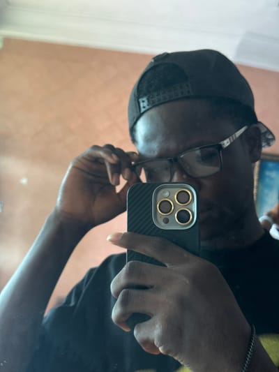 A male individual with dark skin is depicted taking a mirror selfie indoors in Eziobodo, Nigeria. The subject wears a black t-shirt, a black baseball cap reversed, and dark-framed rectangular eyeglasses with silver or white accents on the temples. The subject holds a black smartphone, equipped with a black ribbed case and a visible triple camera array featuring gold lens rings, in their right hand. Their left hand is positioned near the subject's right temple, adjusting the eyeglasses. A thin, silver-colored chain bracelet is present on the subject's left wrist. The background consists of a uniformly painted peach-colored wall. A faint reflection of what appears to be a framed item with a blue hue is visible in the upper right background.