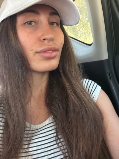 A close-up, medium shot shows a young woman with long, dark brown hair wearing a white baseball cap and a black and white striped t-shirt. She is indoors, likely in the passenger seat of a car, as indicated by the car's interior visible on the right side of the frame and the tinted window with trees visible outside. The woman's head is tilted slightly, and she is looking towards the camera with a neutral expression. Her lips are parted, revealing her teeth. A delicate silver necklace is visible around her neck. The lighting suggests it is daytime, with sunlight filtering in from the window. The background is out of focus, highlighting the subject. There is no specific indication of the location being Larnaca, Cyprus, as the image does not contain any landmarks or contextual elements that would identify the city.