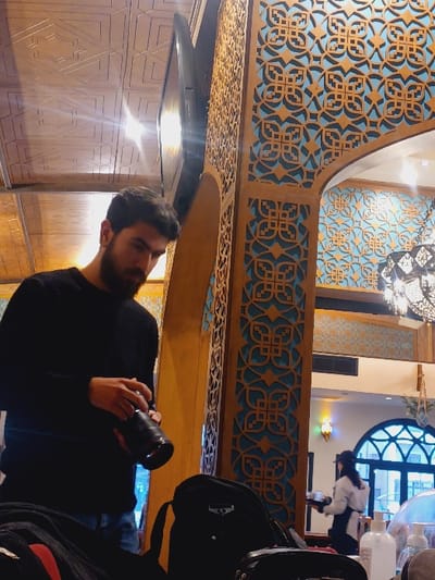 The image depicts the interior of a restaurant or cafe located in Dummar Municipality, Syria. In the foreground, an adult male with dark hair and a beard, wearing a black ribbed long-sleeved sweater and blue jeans, stands holding a black camera with a lens attached, looking down at the device. To his immediate left and right are parts of black bags; one on the left has a visible red strap, and another on the right is a backpack. Several translucent bottles, including one resembling hand sanitizer, are partially visible on a surface to the far right foreground.

The environment features intricate architectural details. The ceiling consists of light wooden panels with geometric patterns. Tall wooden pillars and wall sections are adorned with complex laser-cut or carved geometric latticework in a gold/brown wood tone, backed by a light blue surface. An arched passageway is visible in the mid-ground. A large, ornate metal chandelier hangs from the ceiling in the upper right, and a flat-screen television is mounted high on a pillar in the upper left.

In the background, a female individual, likely a server, is visible. She wears a light-colored long-sleeved shirt, a dark apron, and a dark cap, carrying a tray with items on it in her left hand. Behind her is a large arched window with white framing, offering a partial view of an exterior building or balcony. The lighting suggests an indoor setting with both artificial and natural light sources.