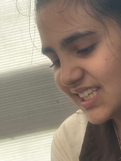 The image is a close-up, profile view of a young person's face, with their head tilted down and to the left. They have dark hair with loose strands framing their face. Their left eye is partially visible and appears to be looking down, while their right eye is obscured. Their mouth is open, revealing white teeth and a hint of pink lips. The person is wearing a light-colored garment, possibly a shirt, with a darker fabric draped over their shoulder, possibly a scarf or the back of a jacket. A thin, gold chain necklace is visible at the bottom right of the image. The background is a blurred, horizontally-striped surface that appears to be a window blind or a similar textured wall. There is no indication of the specific location in Zaliznychne, Ukraine, within the visual information of the image.