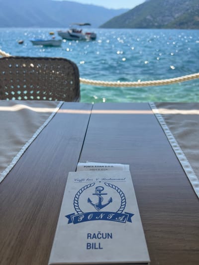 The image shows a table at a cafe or restaurant in Risan, Montenegro, with a view of the bay. In the foreground, a wooden table is set with a beige placemat. On the table, there is a menu or bill from "PONTA RISAN D.O.O." which is identified as a "Caffe bar & Restaurant." The menu/bill features a blue anchor logo and text in both what appears to be a Slavic language ("RAČUN") and English ("BILL"). In the background, the blue water of the bay is visible, with several boats anchored. Mountains form the backdrop, and the water is illuminated by sunlight, creating a shimmering effect. A woven chair is partially visible on the left side of the frame. The scene suggests a relaxed dining experience with a scenic view.