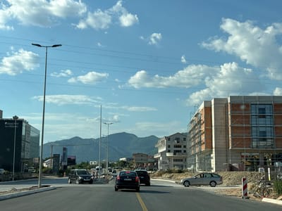 This media file captures a view from a vehicle driving on a road in Radanovići, Montenegro. The scene depicts a developing urban area with ongoing construction, evidenced by buildings covered in scaffolding and piles of building materials. On the left, a modern, dark-glass building with signage for "Hotel Aurel" and "RESTAURANT" stands prominently. In the distance, rolling, tree-covered mountains form a natural backdrop under a bright blue sky dotted with fluffy white clouds.
Several vehicles are visible on the road: a black Volkswagen SUV in the foreground, followed by other cars and a van further down the street. A silver car is parked on the right side of the road near a construction site. Two tall streetlights are positioned along the road, and power lines crisscross the sky.
The time of day appears to be late afternoon or early evening, suggested by the warm light and long shadows. The weather is clear and sunny, with a few clouds contributing to a picturesque sky. The overall impression is one of progress and growth in a scenic, mountainous region.
Visible text includes "Hotel Aurel", "RESTAURANT", and various signs related to businesses and potentially advertisements, such as one for "TOYOTA". License plates are also visible on some vehicles, though the text is difficult to read clearly.