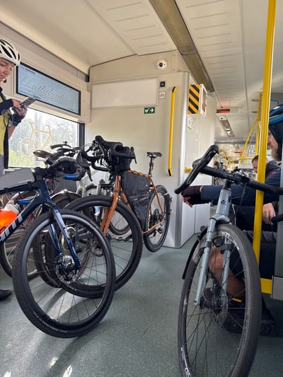 The image depicts the interior of a train carriage with multiple bicycles, suggesting transportation for cyclists. In the foreground, a person wearing a white cycling helmet and yellow jacket is looking at their phone. Several bicycles are parked, some with saddlebags and one with a water bottle attached. The train carriage features yellow handrails, signs indicating "WC" with a bicycle symbol, and a digital display showing "STACJA: Halinów" and "KOŃCOWA STACJA: Siedlce." This confirms the location as Halinów, Poland. The lighting suggests it is daytime, with natural light coming through the windows. The overall scene conveys a group of cyclists using public transportation for their journey.