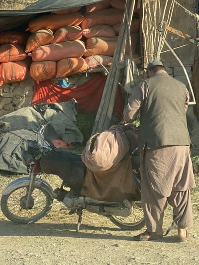 In Arghandab, Afghanistan, an adult male, dressed in traditional clothing with a waistcoat and a small cap, is loading goods onto a motorcycle. The motorcycle, equipped with a side stand, is heavily laden with bundles. The scene takes place next to a structure that is either a makeshift shelter or a building with a canopy. A large pile of bags, possibly containing produce, is visible inside the shelter.
