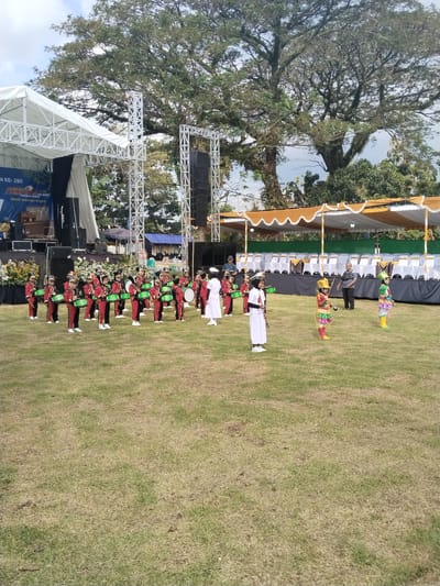 The image captures an outdoor scene in Sambirejo, Indonesia, featuring a marching band performance on a grassy field. The band consists of individuals dressed in red, green, and black uniforms with drums, and others in white uniforms with ceremonial swords. Two individuals in colorful costumes stand apart from the main group.

Behind the band, there is a stage with a white truss structure and speakers. To the right of the stage is a canopy structure with white chairs underneath. In the background, there is a large tree with dense foliage.

The scene appears to be a formal event or celebration. Several adults can be seen overseeing the performance. The weather is clear, with a partly cloudy sky.
