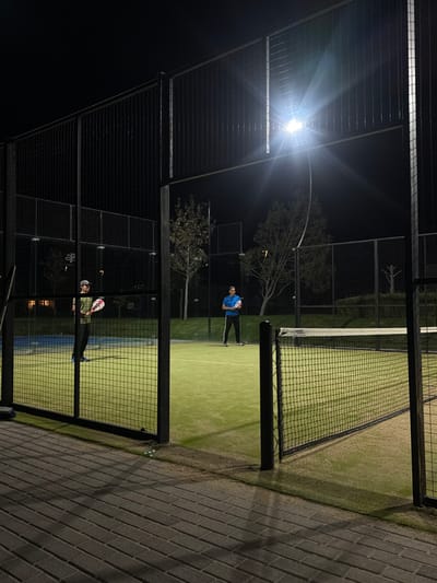 Two people play paddle tennis at night. A bright streetlight illuminates the green court enclosed by a black metal cage. One player holds a tennis racket, ready to hit a ball. A net separates the players. The court borders a brick walkway. Trees are visible beyond the enclosure.