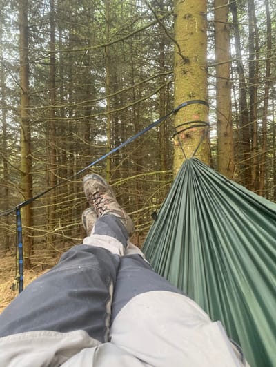 The image shows a person relaxing in a green hammock in a dense forest. The hammock is suspended between two trees, with the person lying back and their feet resting on the ground. The person is wearing grey pants and brown hiking boots. The forest is a mix of evergreen and deciduous trees. The image is taken from a first-person perspective, with the person's feet and legs in the foreground. The location is a forested area in Whaley Bridge, United Kingdom, with a latitude of 53.278 and a longitude of -1.97.