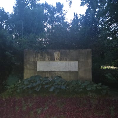 This is a photograph of a memorial monument in the city of Piešťany, Slovakia. The monument is made of concrete and features a relief of a face and the name "BEETHOVEN" inscribed above a rectangular plaque with a relief of musical notes and symbols. The monument is surrounded by lush green foliage, including large-leafed plants at its base. The ground in front of the monument is covered with dark red mulch or ground cover. In the background, dense trees obscure the sky, suggesting the memorial is situated in a park or garden.
