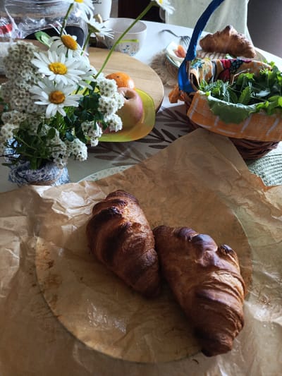 The image depicts a breakfast setting on a table, possibly in Alexandrovskaya, Russia. In the foreground, two croissants are placed on crumpled brown parchment paper. In the background, a bouquet of white daisies and smaller white flowers is in a decorative blue and white patterned vase. To the right of the flowers, a woven basket contains fresh green leaves, possibly herbs or salad greens, lined with a colorful patterned cloth. A blue handle extends from the basket. Further back, a croissant rests on a small white plate with a fork. An orange and an apple are visible on a yellow dish. The overall scene suggests a casual, fresh meal.
