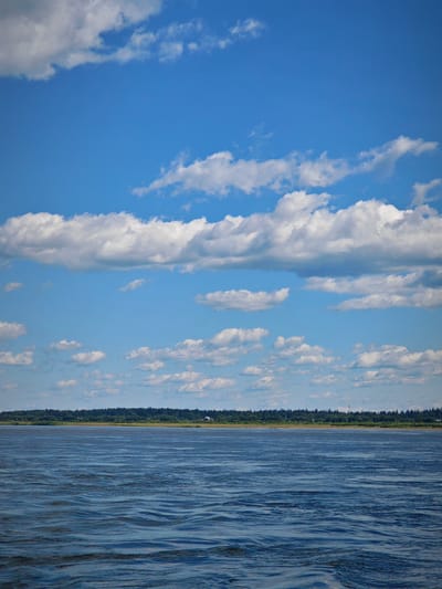 A wide, blue river stretches across the lower half of the image, with visible ripples on its surface. In the distance, a dense green tree line borders the far bank. Above the tree line, a few scattered houses are visible. The upper half of the image is dominated by a bright blue sky with numerous white, fluffy clouds. The scene is peaceful and devoid of any human presence or activity. The location is a riverbank in the village of Абалаково, Russia.