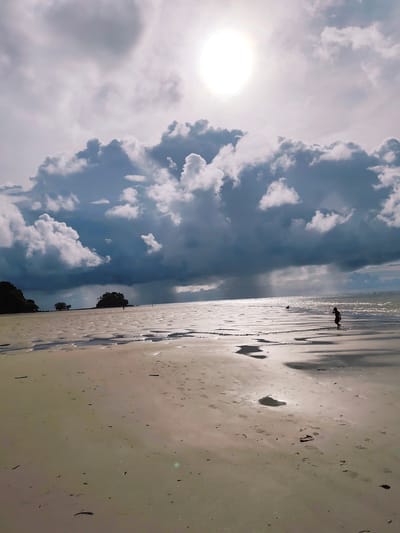 A wide shot captures a beach in Sakhu, Thailand, under a dramatic sky. The sun is a bright, hazy orb in the upper right quadrant, partially obscured by large, dark, and billowing clouds that dominate the upper portion of the frame. The clouds appear to be gathering for a storm, with visible shafts of rain falling in the distance over the sea. The wet sand of the beach reflects the diffused sunlight, creating shimmering patterns. Footprints are visible in the sand, leading towards the water. In the mid-ground, a small island with trees is situated on the left, and further out to sea, two figures are walking on the wet sand. A solitary figure, a child, is in the foreground on the right, silhouetted against the bright water, appearing to walk along the shoreline. The overall atmosphere is one of impending weather change and the vastness of the natural environment.
