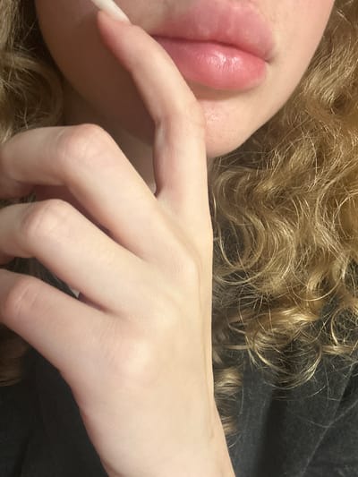 The image is a close-up shot of a person's face and hand. The person has fair skin and blonde, curly hair. Their lips are pink and appear to be slightly glossy. A hand with long fingernails is positioned near their mouth, with fingers touching the lips. The background is dark and out of focus, showing a hint of clothing. There is no visible information in the image that would indicate a location in Vasilyevo, Russia, or any specific urban environment.