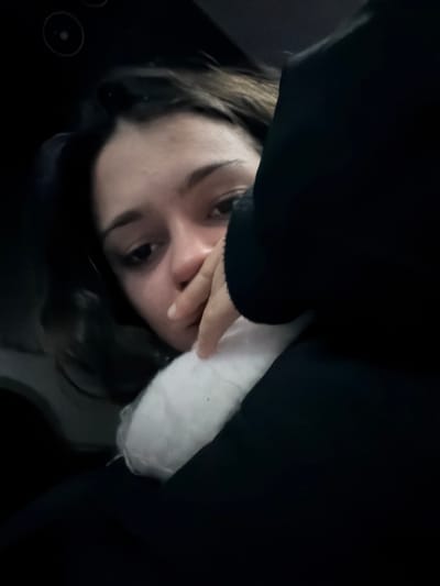 A person lies in bed with a dark, perhaps black, covering or garment close above their face. The individual's face is turned towards the viewer, and their hand is near their mouth. The person has dark hair and light skin. A white pillow or similar cushioning is also visible. The surroundings are shadowy, suggesting a dimly lit room. Part of a dark surface is on the bottom left.