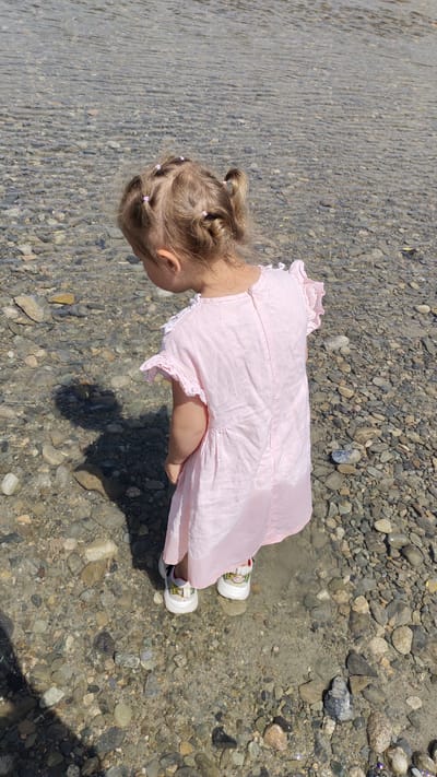 A young girl with blonde hair styled in small pigtails stands in shallow, clear water on a rocky riverbed. She is wearing a pink, ruffled short-sleeved dress and white sneakers with colorful accents. The girl is facing away from the camera, looking down at the water and pebbles. Her shadow is cast on the rocky ground to her left. The background shows more of the rocky riverbed and rippling water, suggesting a natural outdoor setting. This scene could be near the Karatal River, a prominent waterway flowing through or near Taldykorgan, Kazakhstan, as it is known for its shallow sections and rocky shores.