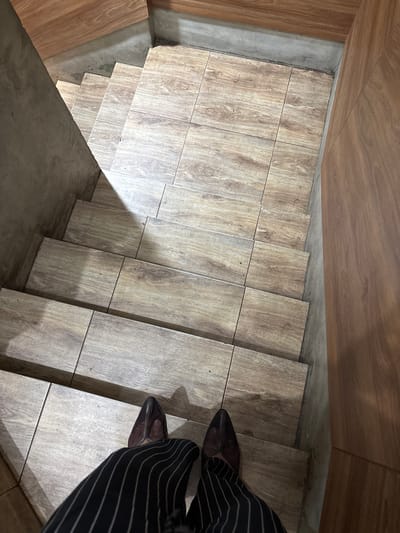 This media file features a close-up, overhead view of a person descending a staircase. The staircase is constructed with large, light-brown tiles that mimic the appearance of wood grain. The walls flanking the staircase are also clad in a similar wood-patterned material, with some sections of exposed concrete visible.
The subject is wearing black trousers with thin white pinstripes and pointed-toe, dark brown shoes, suggesting a formal or semi-formal attire. The lighting suggests an indoor environment, possibly during the day, with shadows cast by the steps indicating the direction of light. The scene appears to be a modern interior, possibly a home, office, or commercial space in South Tangerang, Indonesia. No text is visible in the image. The perspective is from a high angle, looking down the stairs, and the focus is on the steps and the person's feet.