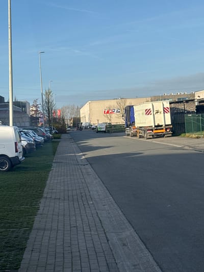 This image depicts an industrial street scene in Roeselare, Belgium, specifically along Oostlaan, facing towards the TRP facility at Oostlaan 460. The environment is an industrial zone under a clear blue sky with minimal cloud cover, suggesting daylight conditions, possibly morning or late afternoon, given the long shadows cast from left to right across the pavement.
In the foreground on the left, a paved pedestrian sidewalk composed of grey interlocking bricks runs parallel to the asphalt road. Adjacent to the sidewalk, a strip of green grass contains a row of parked vehicles, predominantly dark-colored cars and vans, facing away from the viewer. A white panel van is partially visible in the immediate foreground, parked on the grass. A tall, light-colored pole stands vertically on the far left.
The asphalt road, appearing clean, occupies the center and right portions of the frame. A white arrow is painted on the road surface, pointing forward, indicating traffic flow. A blue and white semi-truck is positioned on the road, facing away from the viewer, carrying a large, light-colored rectangular container. The container's rear features orange and white diagonal hazard stripes, a dark symbol (possibly an animal or logo), and the number "15" near the top right.
In the mid-ground, on the right side of the road, a large, light-brown industrial building is visible with a prominent red "TRP" logo on its facade. To the right of this building, a green chain-link fence encloses an area containing stacked dark industrial materials. Further down the road, multiple white vans are visible, some with green markings. Bare trees and red banners are present in the mid-ground, to the left of the TRP building. No individuals are visible within the frame.