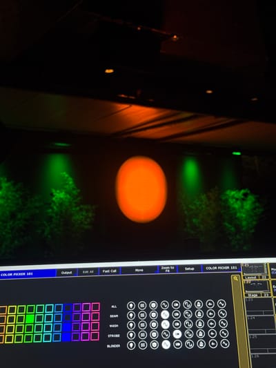 The image displays a stage lighting control interface on a screen, with a blurred backdrop of a stage illuminated by green and orange lights. The stage features what appear to be artificial bushes and a large, circular orange spotlight in the center. The control interface shows a grid of colored squares, likely representing color palettes, and various buttons and icons for light control functions such as "Output," "Edit All," "Fast Call," "Move," "Zoom to Fit," and "Setup." Below the color grid, labels like "ALL," "BEAM," "WASH," "STROBE," and "BLINDER" are visible, accompanied by rows of circular icons. The top right portion of the screen shows layout and stage numbering, with "1.21," "1.22," "1.23," "1.24," "1.25," and "1.26" indicating different lighting zones or fixtures. The image is taken from a low angle, looking upwards towards the stage and then down at the control console. The scene is dark, with the primary illumination coming from the stage lights and the illuminated screen. There are no people or discernable interactions visible in the image. The city of Grumello del Monte, Italy is not identifiable in this image.