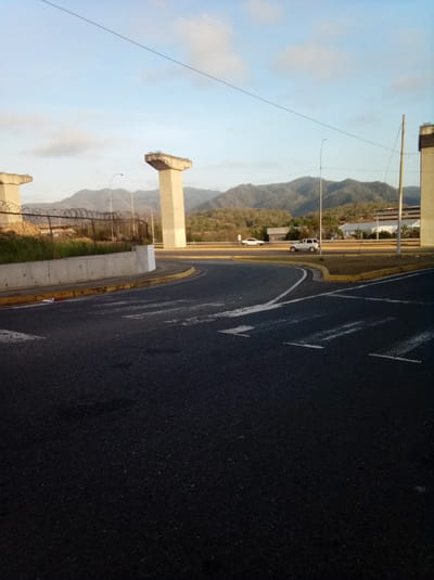 The image shows a street scene in Parroquia Guatire, Venezuela. The road is asphalt with painted white direction arrows and yellow curb markings. There are two concrete pillars, possibly bridge supports, visible. A chain-link fence runs along a low concrete wall near the road. In the background, there are mountains and a building. Two cars are visible in the middle distance. The sky is blue with some clouds. A utility pole with wires is also in view.