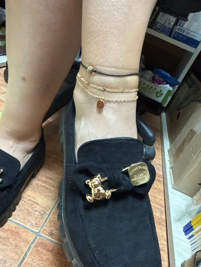 The media file shows a close-up, waist-down view of a person wearing black slip-on shoes with gold accents. The shoes have a soft, textured material, possibly suede or velvet, and are adorned with a gold bow-shaped embellishment that appears to have a logo, and a small gold charm resembling a teddy bear. The person is also wearing delicate gold chain anklets. One anklet has a small, round gold charm. The background is slightly blurred, but suggests an indoor setting with shelving units holding various items, including what appear to be boxes and packages. The floor is tiled with brown and beige tiles. The lighting suggests it is daytime. There is no clear indication of the specific activity taking place, beyond the person being posed or resting. The image is a static shot and does not convey any particular emotion. No legible text is visible in the image. The location context is Aksakovo, Bulgaria.