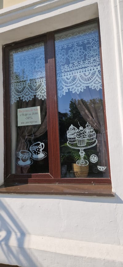 This is a medium shot of a window in Miass, Russia, adorned with white decorative decals. The window frame is dark brown. On the left pane, a sign with Russian text reads "Вкусный час с 18.00 до 20.00 -30% на десерты," which translates to "Happy Hour from 18:00 to 20:00 -30% on desserts." Below the sign, two coffee cups with steam rising from them are depicted. The right pane features a tiered stand with three cupcakes, a potted plant underneath, and two slice-of-lemon decals. White lace-like patterns are applied across the top of both panes of the window. The exterior wall of the building is painted white, and shadows from an unseen object are cast across it.