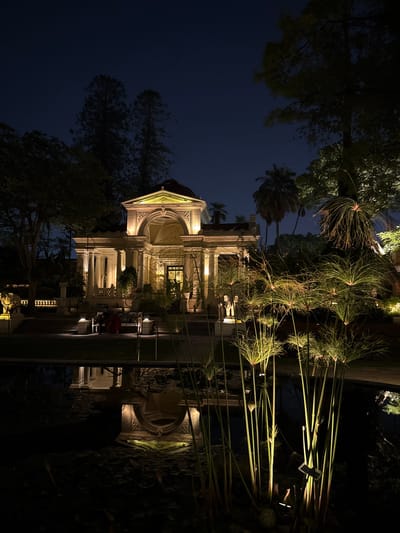 The image shows the illuminated white, ornate building, Kaiser Mahal Library in Kathmandu Metropolitan City, Nepal, at night. The building is two-storied with prominent columns and arched doorways. Sparse trees and plants surround the building. The sky is dark.