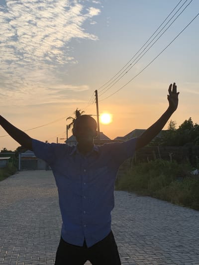 A vibrant outdoor photograph captures a silhouetted individual against a picturesque sunset or sunrise in Ibasa, Nigeria. The image, initially oriented vertically, depicts the horizon running diagonally.

In the foreground, a person, likely male and wearing a striped blue shirt and dark trousers, stands on a paved path. Their arms are expressively outstretched, one reaching high towards the sky and the other extended to the side, creating a pose that suggests joy, freedom, or celebration. The person's head is tilted back, facing the radiant sun.

The sun, a brilliant, low-lying orb, dominates the upper right portion of the scene, backlighting the subject and casting a strong silhouette. The sky transitions from a soft blue with scattered white, wispy clouds on the left to warm golden and orange hues around the sun. Overhead, several utility lines stretch across the sky.

To the right of the person, in the mid-ground, dense green vegetation and some distant structures are partially visible. The paved path, composed of interlocking grey blocks, extends into the distance on the right side of the frame, flanked by more greenery. The warm, golden light of the early morning or late afternoon creates a dramatic contrast and imparts a serene yet energetic atmosphere to the scene. No visible text is present.
