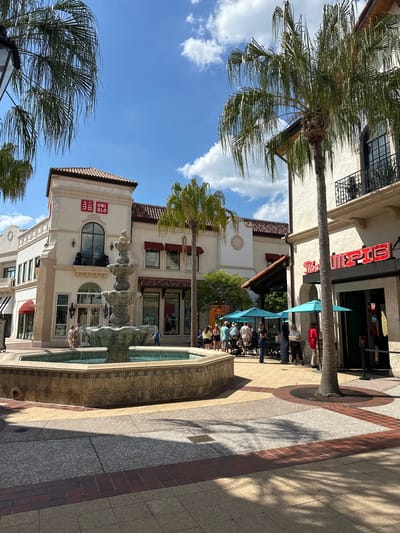 The photograph depicts an outdoor scene in Lake Buena Vista, Florida. A fountain is prominently featured in the foreground, surrounded by patterned tiling. Buildings are visible in the background, including one with a "UNIQLO" sign on its facade. Palm trees are interspersed throughout the scene. People are seated at tables under light-blue umbrellas near a building with "The Daily PIG" in red lettering. The sky is blue with scattered clouds. The shot is taken during daylight, casting shadows across the walkway.