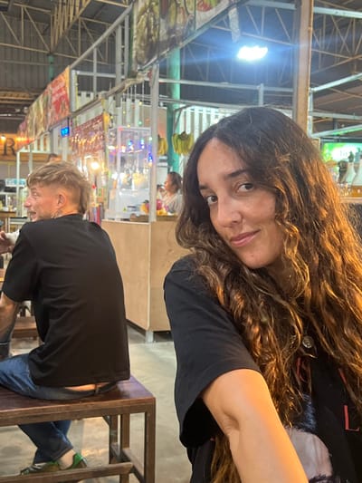 This image depicts a scene within a food court located in Moo 1, Thailand.

In the foreground, on the right, a woman with long, wavy brown hair, wearing a black short-sleeved t-shirt with a graphic design partially visible on the chest, a silver-colored circular pendant around her neck, and a slight smile, is looking directly at the camera. Her right arm is visible.

In the midground, on the left, a man is seated on a dark wooden stool at a matching wooden table, facing away from the camera and looking towards his left. He has short blonde hair styled with a faded undercut and a longer back section. He is wearing a plain black short-sleeved t-shirt and blue jeans, with a portion of his lower back visible above the waistband. A tattoo is visible on his left arm. His left foot, wearing a green shoe, rests beneath the stool.

The background reveals a bustling market or food court environment. Overhead, metal structural beams are visible. Multiple food stalls line the area, featuring large banners displaying images of food. Decorative string lights with small, colored bulbs hang between stalls. A clear display case with illuminated contents stands behind the man. A cluster of green bananas hangs near a stall on the right side of the background. Bottles containing amber-colored liquid are visible on a counter surface further back. Several other individuals are indistinctly visible in the background, partially obscured by the stalls and lighting. The scene is illuminated by artificial overhead lighting and the decorative string lights.