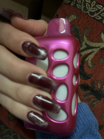 A hand with long, polished burgundy nails is grasping a pink plastic object. The object has a rectangular shape with rounded edges and several circular cutouts revealing a white interior. The hand is positioned against a background of patterned fabric, with a hint of blue fabric visible in the upper right corner. The focus is sharpest on the hand and the object, while the background is slightly blurred.
