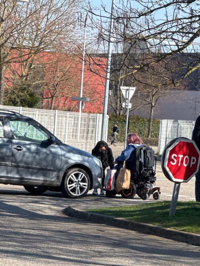 The image captures an outdoor urban scene on a clear, sunny day in Oberhausbergen, France, specifically on Rue des Dreissautwiesen, as indicated by a visible street sign in the background. In the mid-ground, a dark gray five-door hatchback automobile is stationary, positioned with its front left wheel near a curb. Adjacent to the car's front passenger side, a person with dark hair, wearing a black jacket and blue sneakers, is bent forward, appearing to interact with items on the ground. This individual faces a second person seated in an electric wheelchair with orange-accented wheels. The second individual has pink/purple hair and wears a blue jacket. Multiple shopping bags, including a brown paper bag and a striped plastic bag, are affixed to the wheelchair, and another bag is visible on the ground near the first individual's feet. In the immediate foreground on the right, a red octagonal STOP sign with white lettering is mounted on a gray pole. The background features a white metal fence, numerous bare deciduous trees, and a large building with a red facade. A cyclist is visible in the distant background, moving away from the camera. A partial view of a fourth person is present on the far right edge of the frame, next to the STOP sign.