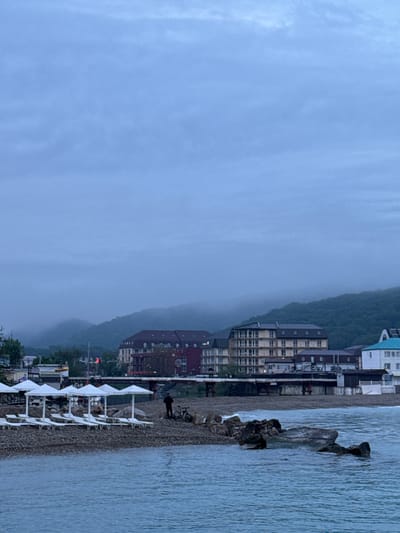 The image depicts a coastal scene in Novomikhaylovsky, Russia. The foreground shows a pebble beach with rows of white sun loungers and umbrellas. A person stands on the beach near a bicycle. In the water, there are some dark rocks. Behind the beach, there are multi-story buildings and a wooded hillside shrouded in fog or low clouds. The sky is overcast, suggesting a gray or rainy day.