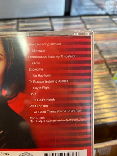 The scene depicts a close-up, angled view of a clear plastic CD jewel case, positioned within what appears to be a retail environment or a large private collection. The location is depicted as being within the city of Edmond, United States.
The primary subject is the back of a music CD, identified by its tracklist as Nelly Furtado's "Loose" album. The CD booklet/cover, visible through the clear case, features a red background with horizontal darker red stripes. On the left side of the cover, a partial image of a person's face, specifically the right eye and lower facial features of what appears to be a light-skinned female with dark hair, is present.
The visible tracklist, printed in white sans-serif font, includes:
* Afraid featuring Attitude
* Maneater
* Promiscuous featuring Timbaland
* Glow
* Showtime
* No Hay Igual
* Te Busque featuring Juanes
* Say It Right
* Do It
* In God's Hands
* Wait For You
* All Good Things (Come To An End)
* Bonus Track: Te Busque (Spanish Version) featuring Juanes
At the bottom left of the CD case, partially obscured text reads "Piracy," accompanied by a fragment of a design resembling an anti-piracy warning label, next to a partial barcode. The plastic jewel case exhibits minor surface scratches and a small chip or imperfection on its top right edge.
The background, out of focus, consists of numerous other CD jewel cases arranged in horizontal rows, indicative of shelving units in a music store or a extensive personal library. No individuals are visible within the depicted scene itself, only the partial image on the album cover. There are no discernible actions or interactions occurring within the frame.