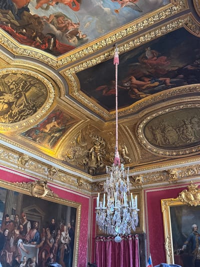 This is a photograph of the interior of a room in the Palace of Versailles, France. The room features ornate gilded ceilings with large paintings depicting mythological or historical scenes. A grand crystal chandelier hangs from the ceiling, illuminated by several candles. The walls are adorned with large framed paintings, including a scene of what appears to be a biblical feast on the left and a portrait of a man in regal attire on the right. The walls themselves are covered in a rich red fabric, possibly damask, with gilded decorative moldings running along the top. Red velvet curtains are visible, framing the paintings and adding to the opulent decor. The overall impression is one of extreme luxury and grandeur characteristic of the Palace of Versailles.