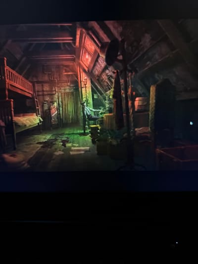 The image depicts a dimly lit, cluttered attic room with a sloped ceiling. A bunk bed with a ladder is positioned on the left side of the room, with the lower bunk made. The floor appears to be wood and has dark splatters, suggesting a possible incident. In the center of the room, a figure wearing overalls and suspenders is seated in a chair at a table filled with items, illuminated by a green light. A large lamp with a bell-shaped shade stands to the right of the figure, and various boxes, barrels, and draped fabric are scattered throughout the space. A tall, stone-like object stands near the right edge of the frame. The overall atmosphere is one of disarray and a somber, perhaps even sinister, mood. The location is not specified to be in Vancouver, United States of America.