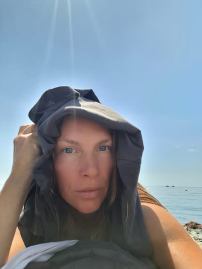 An outdoor, medium shot selfie features a woman with light brown hair and blue eyes, partially obscured by a dark gray hooded shirt, which she is holding up to cover her head and face. She is looking directly at the camera, with a neutral expression. The background shows a clear, bright blue sky with sun flare effects, and a calm, blue sea with two small boats and a buoy in the distance. The foreground includes a rocky shoreline. The image appears to have been taken on a sunny day. The location is identified as Bol'shoi Utrish, Russia.