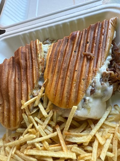 A close-up shot depicts a sandwich with french-fried potatoes, contained within a white, molded, biodegradable-looking container. The sandwich bread is toasted with raised grill marks and appears to be filled with meat and cheese. The french fries are short and slender. The image was taken in Davie, United States of America.