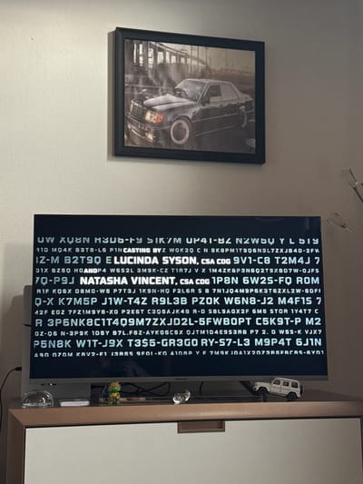 The image shows a room with a framed black-and-white picture of a car hanging on a white wall, next to a TV displaying strings of uppercase letters and numbers. A miniature white G-Wagon model car is positioned on top of the TV, and a small toy figure is visible near the TV's edge. The TV is placed on a wooden cabinet. The location in the city of Katwijk, Netherlands, is not discernible from the image.