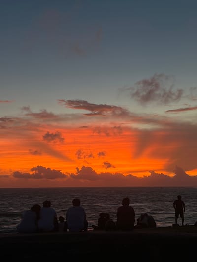 This image captures a sunset scene with silhouetted figures against a vibrant, orange-hued sky. The sky is filled with dramatic clouds, some appearing wispy and light, while others are darker and more defined, stretching across the frame. The sun appears to be setting over the ocean, creating a warm glow.
Several people are visible in silhouette along the edge of what seems to be a pier or a seawall. They are facing the ocean and the sunset. Some are standing, while others are seated. One figure in the distance stands on the seawall, looking out towards the water. The water itself is dark and choppy, reflecting the colors of the sky.
The overall mood of the image is serene and contemplative, with the dramatic colors of the sunset and the quiet presence of the figures suggesting a moment of reflection or enjoyment of nature. The time of day is clearly sunset. There is no visible text in the image. Given the location context, this scene is likely in Galle, Sri Lanka.