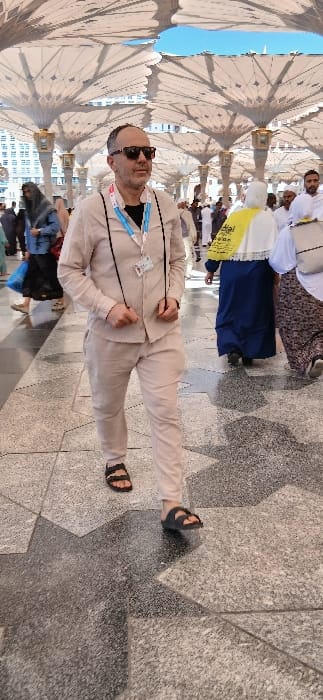 The image depicts an outdoor pedestrian area characterized by large, white, retractable umbrella structures providing shade. This environment is consistent with the courtyards surrounding the Prophet's Mosque (Al-Masjid an-Nabawi) in Medina, Saudi Arabia.

In the foreground, a male individual walks towards the viewer's left. He is of middle age, has short dark hair, stubble, and wears dark sunglasses. His attire consists of a light beige/tan long-sleeved button-up shirt worn over a black t-shirt, matching beige/tan trousers, and dark sandals. A white lanyard with an identification badge hangs around his neck, resting on his chest. A black cord, possibly for an electronic device, also hangs around his neck, with its ends held loosely in his hands. His left foot is lifted mid-stride.

In the background, numerous other individuals are visible, also walking. One woman on the right wears a dark blue dress or abaya, a white head covering, and carries a bright yellow item, possibly a bag or shawl, with dark text partially legible as "Hajj" or similar. Other figures include men in white attire and women in various head coverings and garments. The ground is paved with large, textured granite tiles in light grey and dark grey/black, arranged in a geometric, star-like pattern. Patches of clear blue sky are visible through openings in the umbrella canopy. Distant buildings are discernible in the far background.