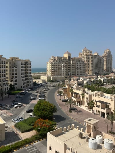 The image depicts a view of a coastal city with modern architecture and a clear blue sky, suggesting a sunny day. The scene is dominated by a cluster of tall, multi-story buildings with light-colored facades, many featuring balconies and arched windows. Some buildings have rounded tops, possibly with domes. In the foreground, a paved road winds through the buildings, lined with parked cars, palm trees, and manicured bushes. Several white water tanks are visible on the rooftops of some of the lower structures. In the distance, the blue ocean meets the horizon, with a sandy area and smaller buildings visible along the coast. The perspective suggests the photo was taken from an elevated viewpoint, looking down on the city and the road. The overall impression is one of a well-developed urban area in a warm climate.