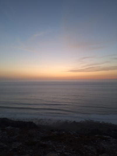 The image depicts a serene seascape at dusk, likely captured from a coastal vantage point in Ahrara, Morocco. The sky transitions from a pale blue at the top to soft hues of orange and pink near the horizon, suggesting the sun has recently set or is about to rise. A few wispy clouds are visible in the upper part of the sky. Below the sky, the ocean stretches out, appearing calm with gentle waves creating subtle ripples. The water is a muted grey-blue, reflecting the twilight colors. In the foreground, the image shows a dark, rocky terrain, characteristic of a rugged coastline. This rocky area is sparsely covered with what appear to be low-lying vegetation or scrub. The overall atmosphere is tranquil and undisturbed, with no people or man-made structures readily apparent, emphasizing the natural beauty of the coastal landscape.