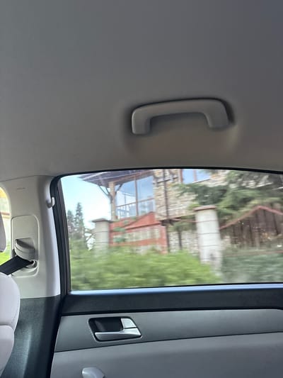 The image depicts a view from inside a vehicle, looking out the window towards a building and vegetation. The interior of the car shows a grey headliner with a grab handle, and a portion of a seat and seatbelt mechanism on the left. The window frames the scene, with a dark trim along the top and bottom. Outside the window, a building constructed with stone and reddish-brown elements is visible, surrounded by green foliage. The scene appears to be taken from a moving vehicle, as evidenced by the slight blur of the exterior. The location is identified as Slaveevo, Bulgaria.
