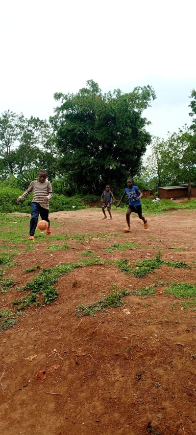 The scene depicts three young males playing football outdoors in Kundu 1, Democratic Republic of the Congo, under an overcast sky. The ground is a reddish-brown dirt field with scattered patches of green vegetation.

In the foreground, slightly left of center, a young male is captured mid-action, kicking a worn, reddish-brown spherical object resembling a football with his right foot. He is wearing a long-sleeved horizontally striped light and dark shirt, dark long trousers, and orange flip-flops. His body is angled towards the right side of the frame.

To his right, in the mid-ground, a second young male runs towards the right. He wears a blue short-sleeved t-shirt with partially visible white text, "how cute," and dark shorts. He is barefoot.

Further back and slightly to the left of the second male, a third young male is visible, facing towards the right with bent knees, indicating movement. He wears a dark t-shirt and patterned shorts. He is also barefoot.

The background features dense green foliage from various trees. On the far right, behind the players, a partially obscured structure made of reddish-brown bricks or mud is visible, along with another smaller, similar formation or pile of materials.