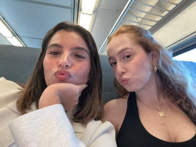Two young adult females are depicted in a selfie taken inside a passenger train or public transport vehicle located in Linden, United States. The female on the left, with shoulder-length dark brown hair, wears a light-colored collared top. She supports her chin with her right hand and purses her lips into a pout while looking directly at the camera. The female on the right, with long, wavy reddish-brown hair, wears a black halter-neck top accessorized with multiple gold necklaces, one featuring an 'E' pendant, and gold hoop earrings. She also exhibits a pouting facial expression and looks towards the camera. The background consists of grey upholstered seating, overhead white luggage racks, white ceiling panels, and a window visible on the far right.