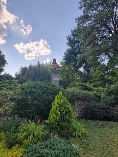 The image shows a statue of a king wearing a crown and holding a scepter, positioned on a pedestal in a lush garden. The statue is made of bronze and is weathered, with a patina of age. The garden is filled with various types of greenery, including bushes, trees, and flowering plants. In the background, there is a stone wall and a clear blue sky with some white clouds. The scene is peaceful and serene, suggesting a historical or memorial site. The statue is located in the city of Hagimus, Moldova.