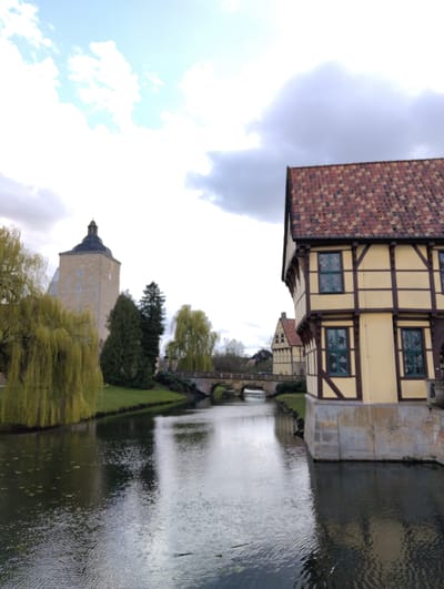 The image displays a scene along a waterway in Burgsteinfurt, Germany. In the foreground on the right, a two-story half-timbered building features light yellow infill panels, dark brown timber framing, and a multi-toned reddish-brown tiled roof. Multiple rectangular windows with dark green frames are visible. The building's base is constructed from light-colored stone and directly borders the water. The water surface shows ripples and reflections of the building and sky.
On the left, a large weeping willow tree with light green foliage droops towards the water. Beyond the tree, a light-colored, square stone tower, identified as part of Steinfurt Castle, rises prominently. The tower has a dark, domed roof with a small spire and features a clock face and several small rectangular windows. Dark green coniferous trees are situated on the grassy bank between the willow tree and the tower.
In the midground, a multi-arch stone bridge spans the waterway. Further along the right bank, past the bridge, another half-timbered building, stylistically consistent with the foreground structure, is partially visible. The sky above is overcast with a mix of bright and darker clouds. No individuals are present in the scene.