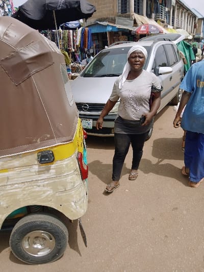 The image depicts an outdoor street scene within a commercial area of Kwekwe, Zimbabwe.
In the foreground, the rear section of a three-wheeled vehicle, specifically an auto-rickshaw with a faded yellow body and a brown canvas roof, is visible on the left. Its rear wheel, with a silver hubcap and black tire, is prominent, along with a black mudflap behind it.
In the midground, an adult female is walking right-to-left across the frame. She wears a light grey short-sleeved top with dark markings, dark grey leggings, and light-colored flip-flops. A white fabric head covering is on her head, and she holds a dark object in her left hand. A dark band is visible on her upper right arm. Directly behind her is a silver minivan, possibly an Opel Zafira, with its front visible and a partial view of its license plate showing "KGE 1ZZME." The vehicle's rear hatch or trunk appears to be slightly open. To the immediate right of the central female, another individual's torso and legs are visible, wearing a blue t-shirt and blue trousers, also walking towards the right.
The background features multiple market stalls and structures lining the street. A large black umbrella, partially obscuring a "NIVEA" sign, covers a stall displaying clothing. Further back, another stall is visible with an "Airtel" branded red umbrella. Buildings with upper floors, windows, and balconies are present, suggesting multi-story commercial or residential structures. Goods, predominantly clothing, are hung for sale. The ground surface is a light brown, dusty material, consistent with an unpaved or worn road. Shadows suggest direct overhead sunlight.