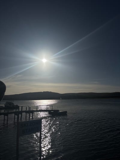 The image captures a vibrant, sun-drenched waterscape, likely in Port Alfred, South Africa. A brilliant sun is positioned on the left side of the frame, emitting a strong lens flare and bright sunbeams across the sky. Its intense light creates dazzling, shimmering reflections on the surface of the water, which occupies the right half of the image and stretches into the distance. The water appears to be rippling gently, catching the sunlight.

In the mid-ground, a dark, undulating silhouette of distant land or hills spans the horizon, creating a stark contrast against the bright sky and glistening water. Extending from the right edge into the water is a wooden dock or jetty, largely rendered in silhouette due to the strong backlighting. Visible are several pilings and horizontal supports, with their shapes subtly reflected in the water beneath.

A white sign with black capital letters is prominently displayed on the dock in the lower right quadrant. The text clearly reads: "SKI ZONE STARTS".

The scene suggests a bright morning or late afternoon, with the sun relatively low in the sky, causing dramatic backlighting. The clear sky and sparkling water indicate fair weather, and the overall atmosphere is one of serenity and warmth. No people are explicitly visible within the frame, allowing the focus to remain on the natural beauty and the subtle hint of recreational activity implied by the sign.