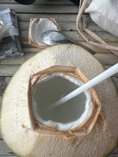 An opened green coconut, cut in half, reveals its translucent water and white flesh. A white straw protrudes from the liquid. Next to it, a silver spoon rests on a piece of coconut shell, partially filled with coconut meat. A package labeled "Bali Shag White Halfzware" is partially visible. A cloth bag with tan straps sits in the top left corner. The composition is shot from above on what appears to be a wooden surface.